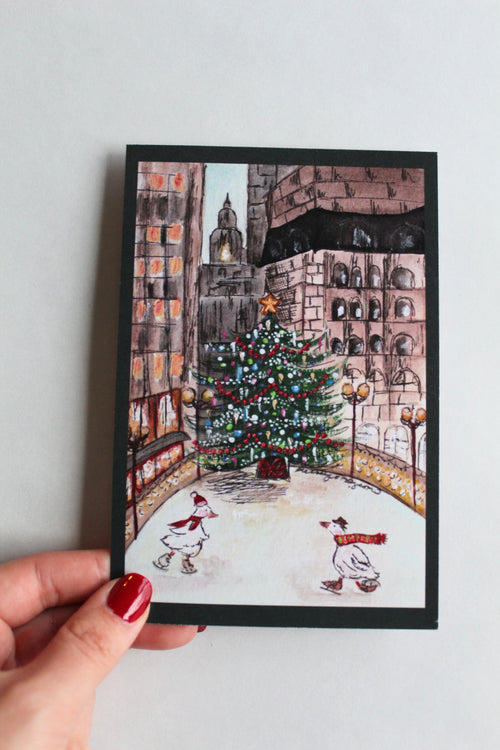 Postcard with an illustration of two ducks skating in front of a Christmas tree, wearing hats and scarves, in front of a city landscape. 