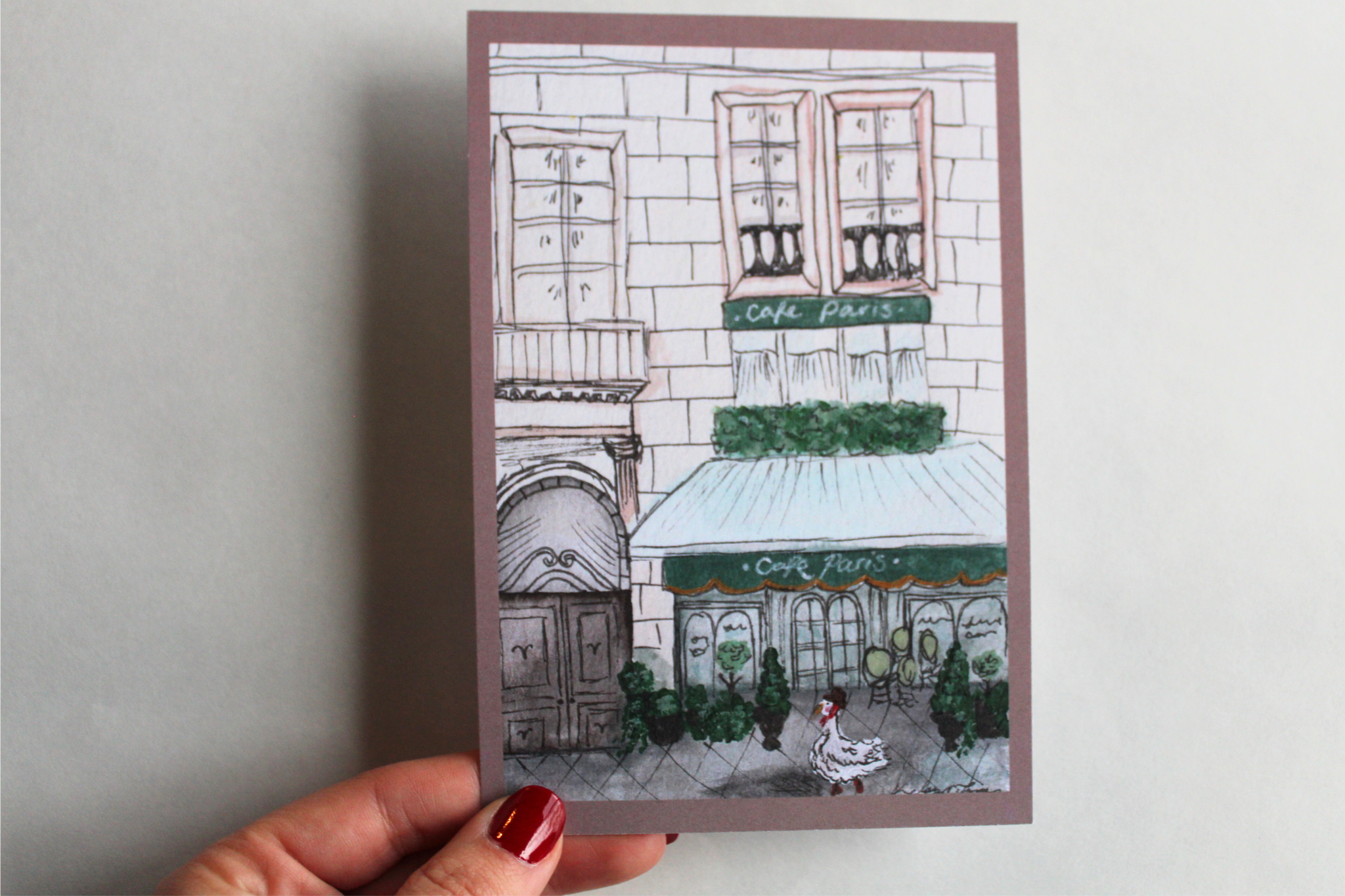 Postcard with an illustration of a duck in Paris, walking down the street wearing a scarf and hat in front of a large building with a cafe attached. 