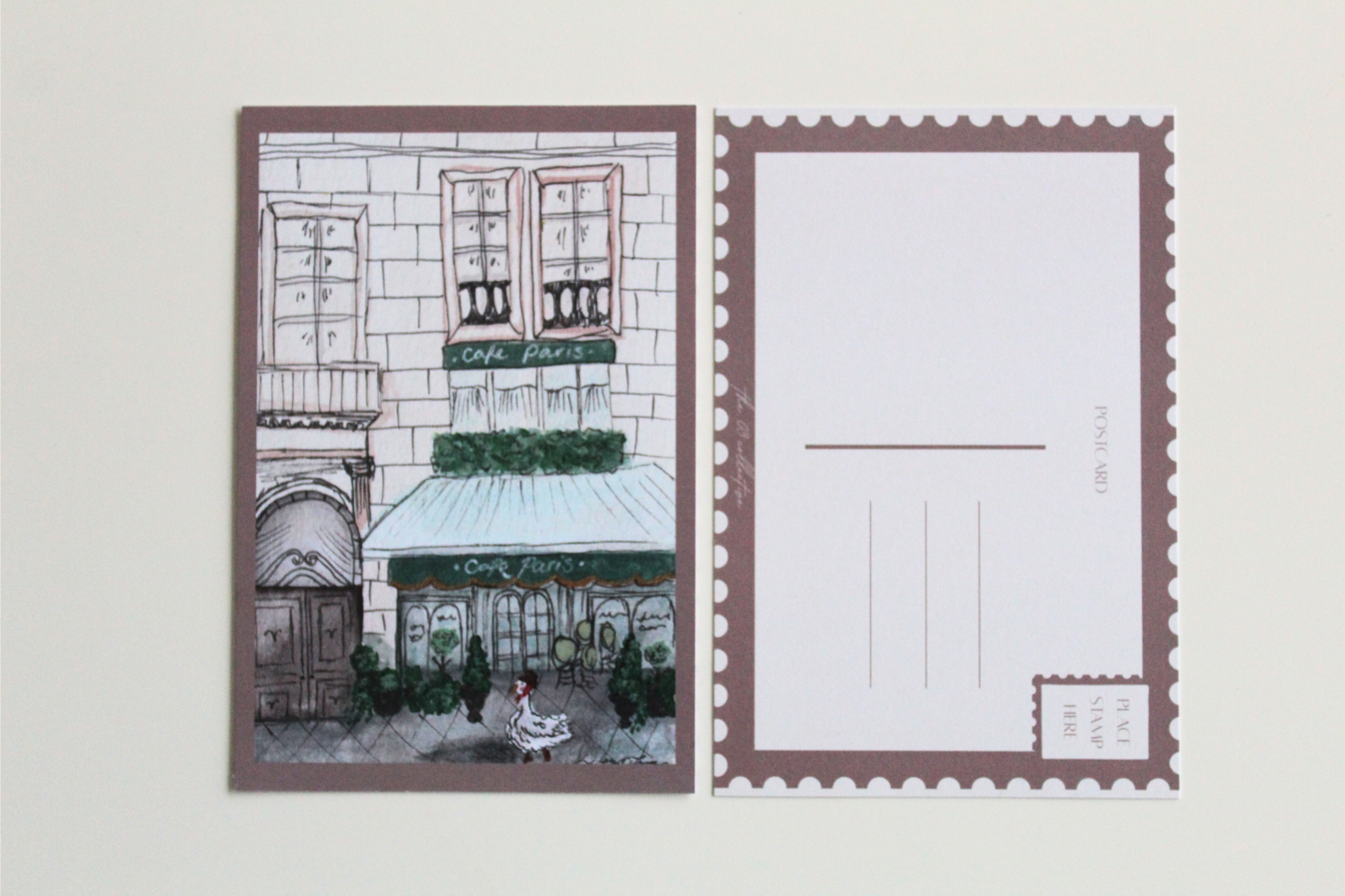 Postcard with an illustration of a duck in Paris, walking down the street wearing a scarf and hat in front of a large building with a cafe attached. Pictured with the back of the postcard where a letter can be written and address lines.